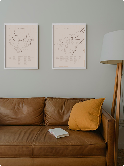 Two framed Elevated Trail Maps posters, Mt. Fromme and Mt. Seymour, hanging above a tan leather sofa beside a wooden floor lamp